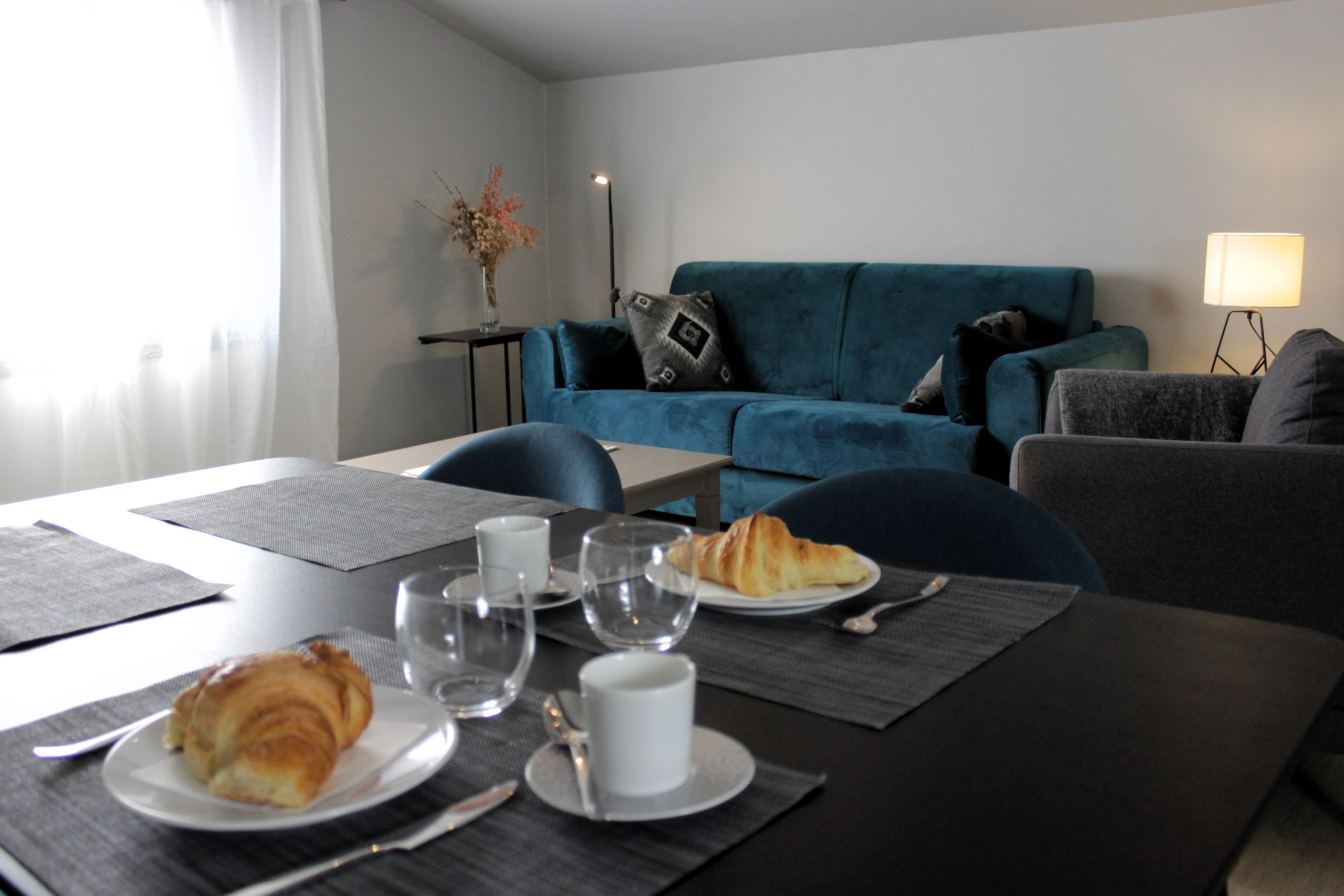 Une pièce lumineuse et moderne, décorée dans un style sobre et élégant. Au premier plan, une table noire est dressée pour le petit-déjeuner avec deux assiettes contenant des croissants, des verres ronds et des tasses blanches sur des sets de table gris. À l’arrière-plan, un canapé en velours bleu turquoise est agrémenté de coussins à motifs géométriques noirs et gris. À droite, une lampe de table diffuse une lumière douce, et un bouquet de fleurs séchées décore une table basse. L’ensemble crée une atmosphère accueillante et cosy.