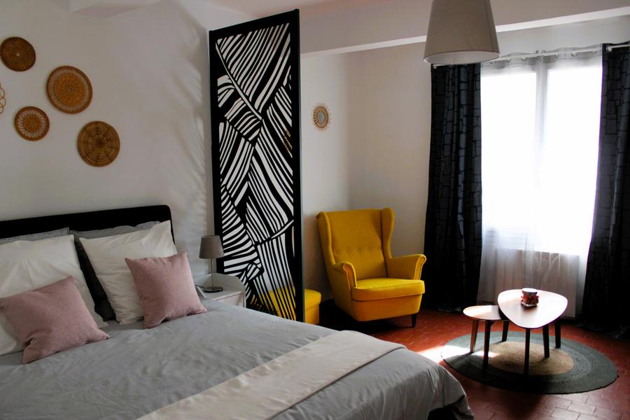 chambre blanche avec lit 2 places avec paravent moderne noir, fauteuil jaune et petites tables basses sur tapis rond vert et beige