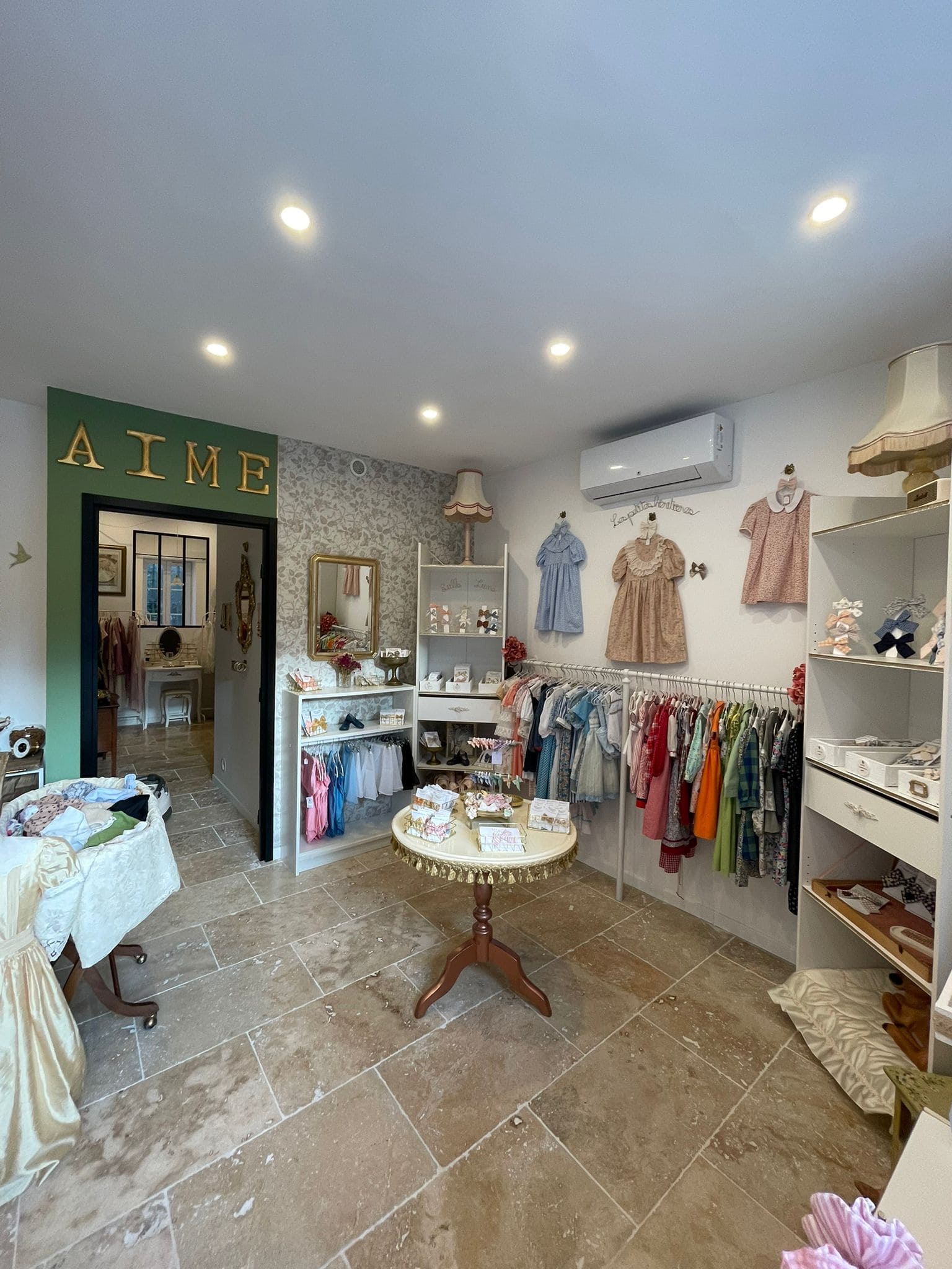 Un espace boutique lumineux avec un sol en pierre claire et des murs blancs décorés. Plusieurs vêtements pour enfants sont accrochés à des cintres : petites robes, chemises et ensembles colorés. À droite, un portant présente une série de tenues variées. Sur la gauche, une table ronde recouverte de produits (probablement accessoires ou petits articles de mode). L’ambiance est douce et élégante, avec des détails rétro comme une lampe à abat-jour en tissu et des lettres dorées formant le mot AIME au-dessus de la porte.