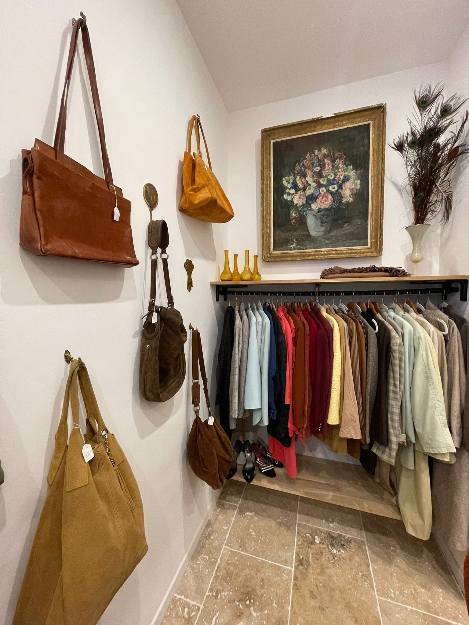Une petite pièce chaleureuse dédiée à la mode féminine. Des sacs en cuir et en daim sont suspendus au mur à gauche. Sur le portant, une collection de vestes, manteaux et chemises colorées dans des tons sobres et chauds : beige, rouge, bleu, vert. Des chaussures à talons sont posées en dessous. Au mur, un grand tableau encadré représente un bouquet de fleurs dans un vase, à côté d’un vase avec des plumes de paon et de petites carafes jaunes. L’atmosphère rappelle une boutique de seconde main chic et soignée.