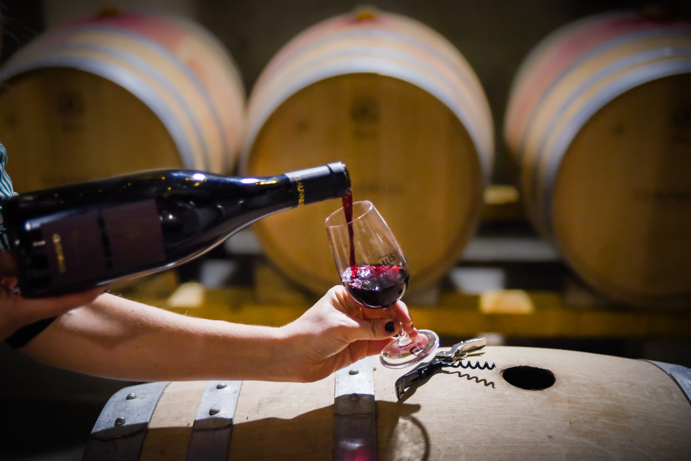 Un verre de vin rouge Clauvallis est servi depuis une bouteille, posé sur un tonneau en bois dans une cave. En arrière-plan, plusieurs barriques de chêne sont alignées, créant une ambiance chaleureuse et authentique de caveau de dégustation.
