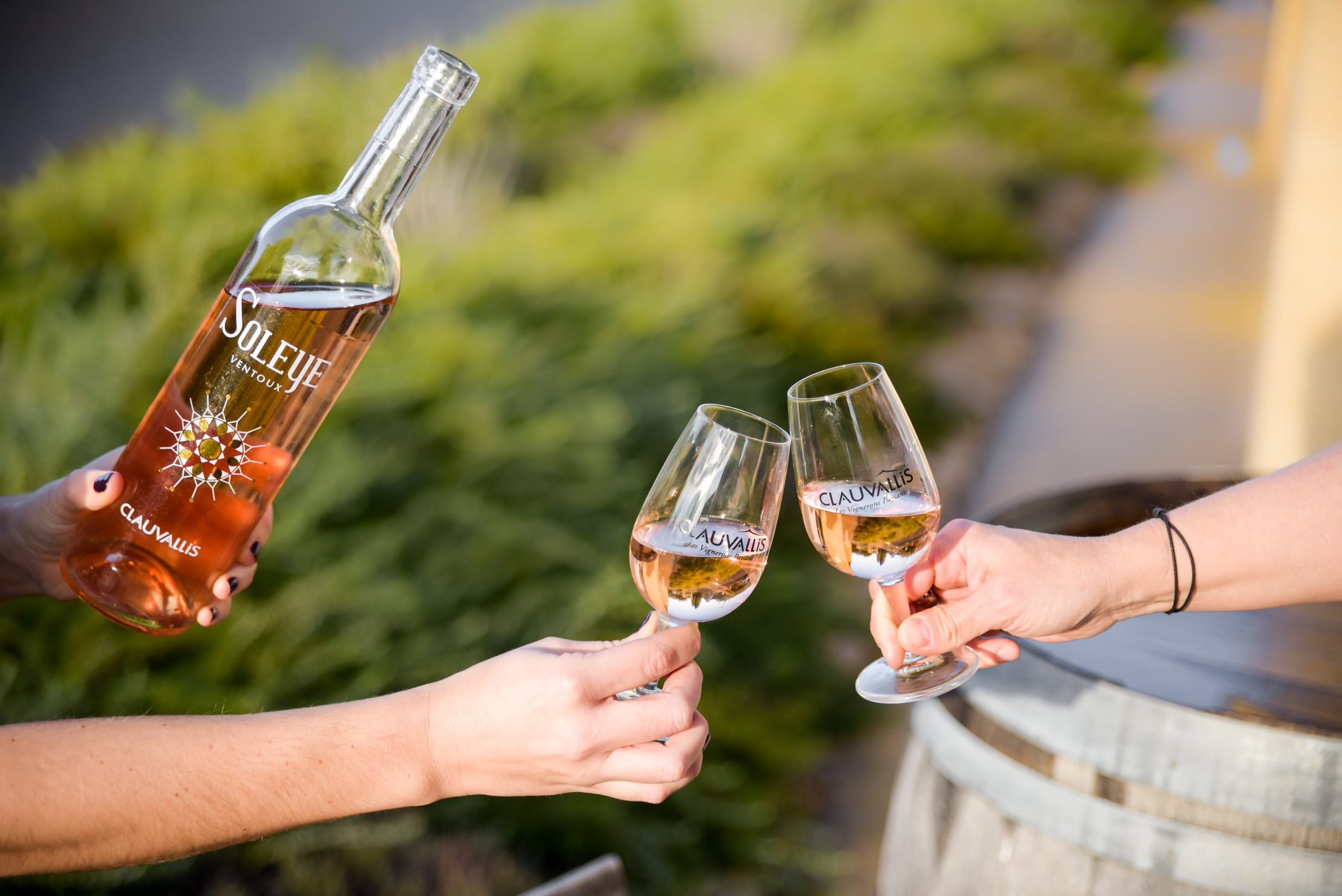 Deux personnes trinquent avec des verres de vin rosé Clauvallis, devant un tonneau en bois et des buissons verts en arrière-plan. Une main tient la bouteille de rosé "Soleye Ventoux", posée légèrement en avant de la scène.
