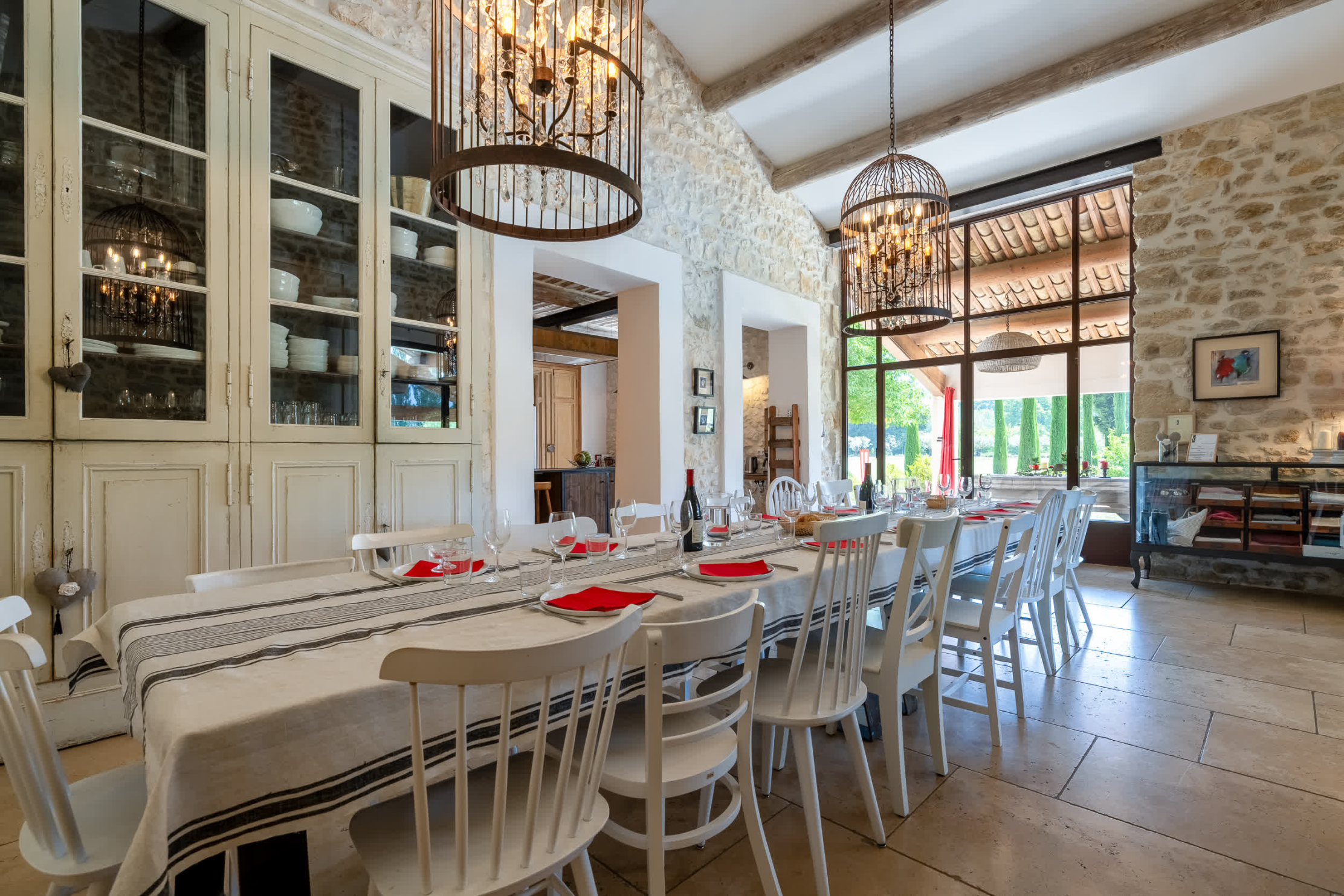 Grande salle à manger conviviale dans une bâtisse en pierre, avec longue table dressée, lustres élégants et larges ouvertures sur l’extérieur.