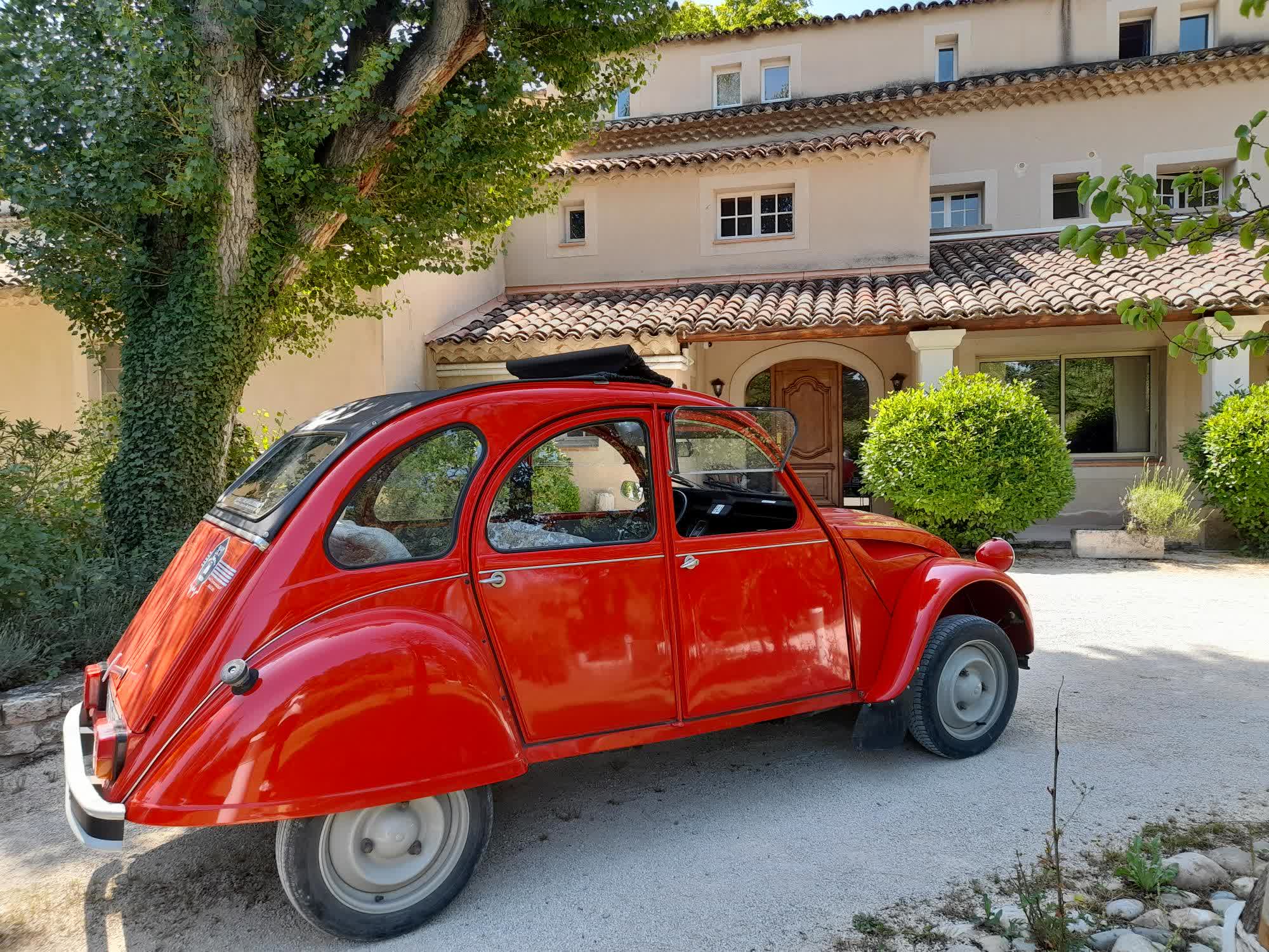 Une voiture ancienne Citroën 2CV rouge vif est garée devant une grande maison provençale aux murs clairs et au toit en tuiles, entourée d’arbres et de verdure.