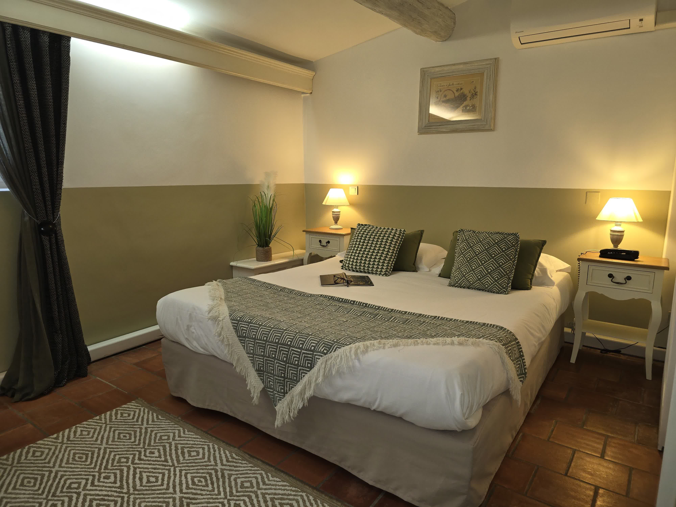 Une chambre double décorée dans un style provençal avec des poutres apparentes et un sol en terre cuite, équipée d’un grand lit recouvert de coussins verts à motifs géométriques, de deux tables de chevet avec lampes et d’un cadre accroché au mur.