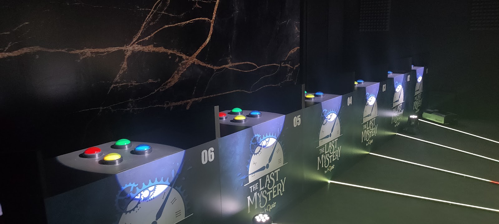 La photo montre plusieurs pupitres numérotés, alignés dans une salle sombre. Chaque pupitre possède quatre boutons lumineux de couleurs différentes : rouge, vert, jaune et bleu. Le décor, intitulé « The Last Mystery », représente une horloge et un œil mécanique. Des bandes lumineuses au sol créent une ambiance mystérieuse.