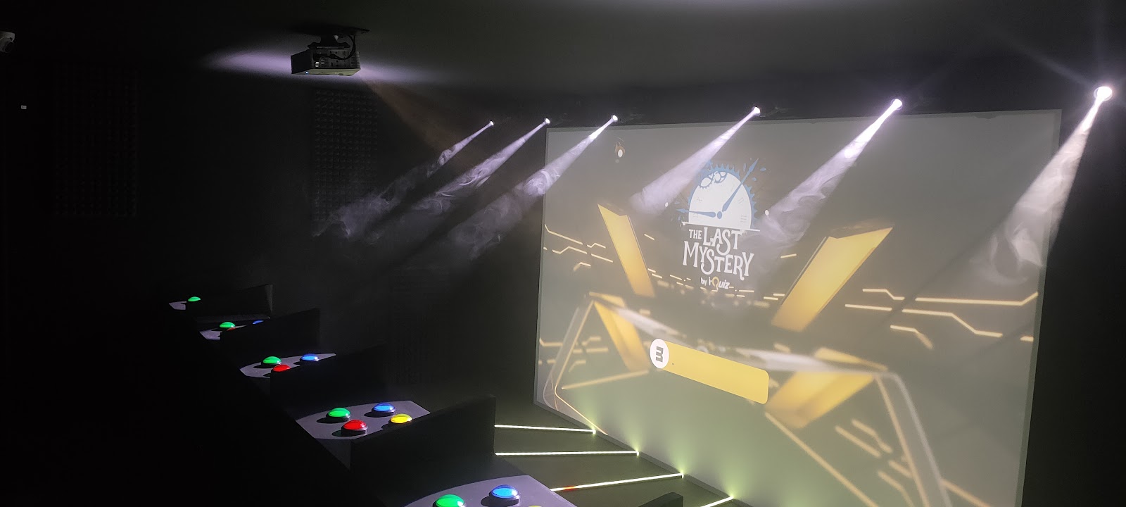 On découvre la même salle de jeu sous un autre angle. Plusieurs projecteurs éclairent un grand écran sur lequel apparaît le titre « The Last Mystery ». Les pupitres avec leurs boutons colorés sont placés face à l’écran. La fumée, les faisceaux lumineux et l’obscurité donnent une atmosphère immersive, semblable à celle d’un jeu télévisé ou d’un escape game.