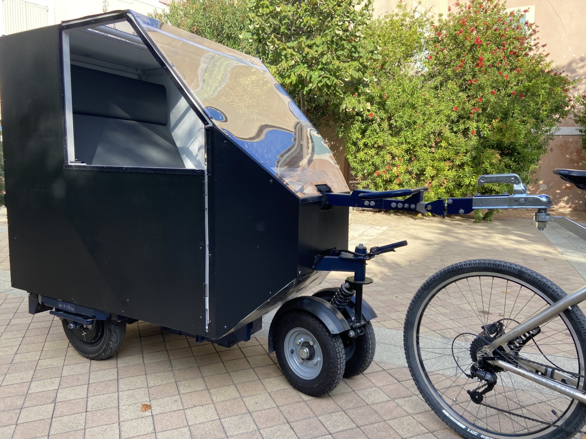 Vélocab noir à trois roues, attelé à un vélo, stationné en journée sur une placette pavée avec des buissons fleuris en arrière-plan.