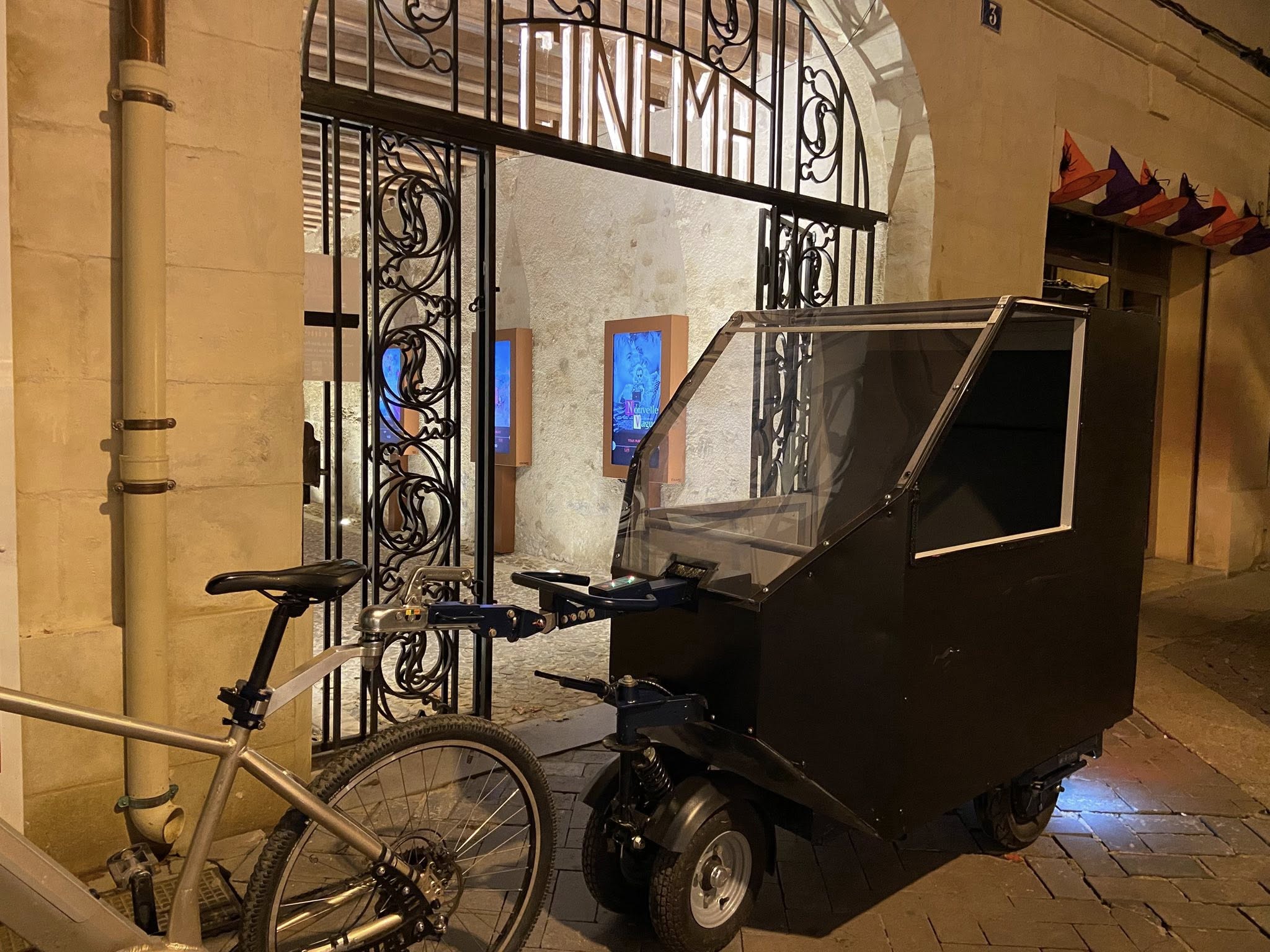 Un vélocab noir stationné de nuit devant l’entrée d’un cinéma ancien, éclairé et encadré par une grille en fer forgé.