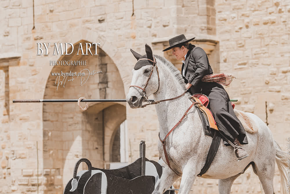 Un cavalier en habit traditionnel monte un cheval gris clair et vise une cible avec une lance devant un bâtiment en pierre.