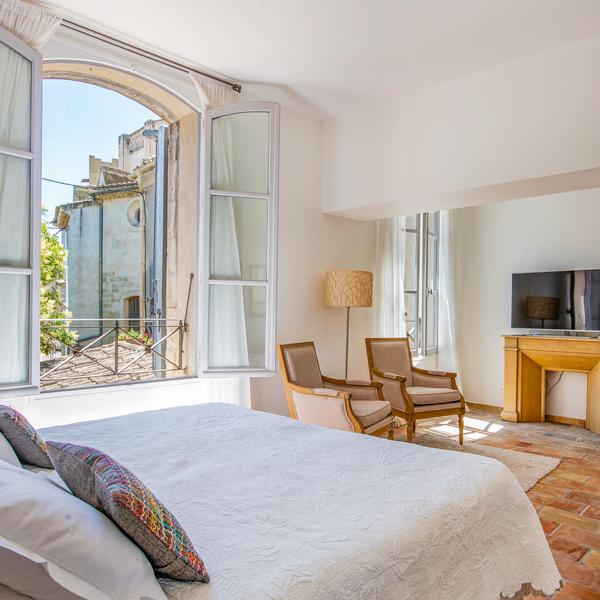 grande chambre en parfeuille avec fenêtre donnant sur les toits de la ville. manteau de cheminée jaune ocre et grand lit au premier plan avec dessus de lit en boutis blanc