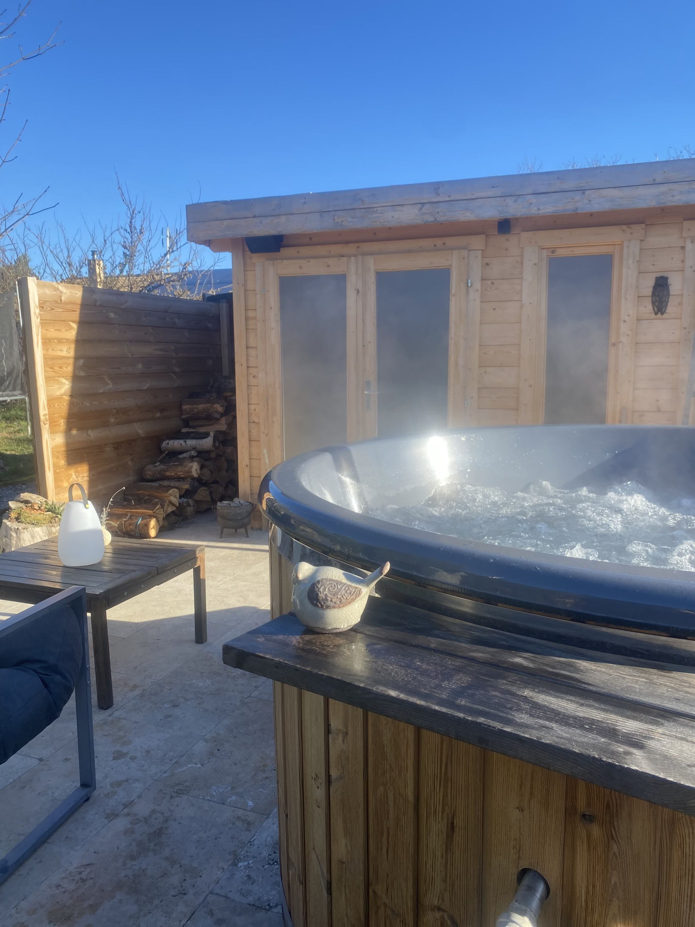 Bain nordique extérieur en bois, chauffé et fumant, installé sur une terrasse ensoleillée devant un chalet en bois.