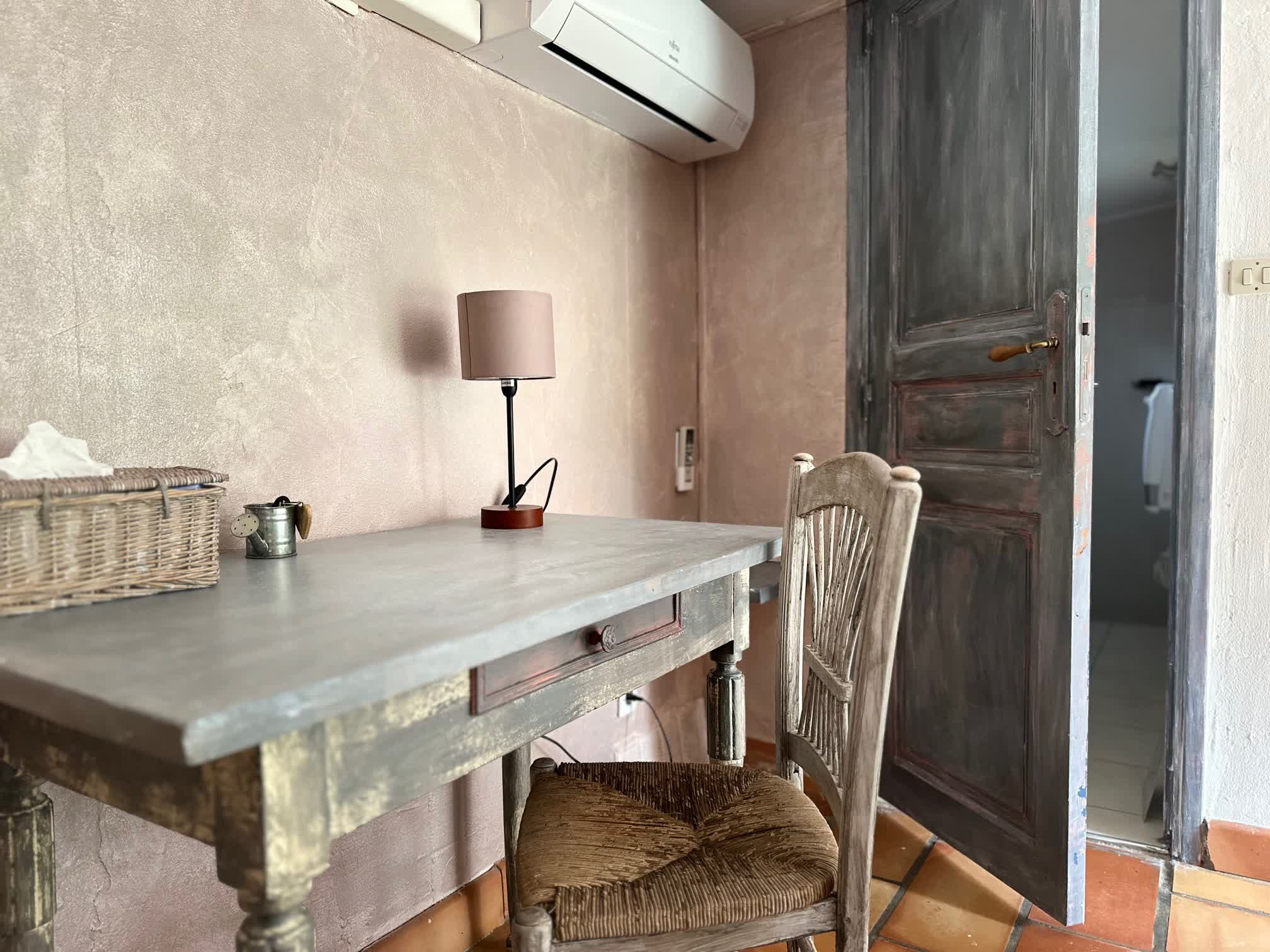 Coin bureau en bois patiné avec chaise et lampe, situé dans une chambre aux tons doux, à côté d’une porte ancienne menant à la salle d’eau.