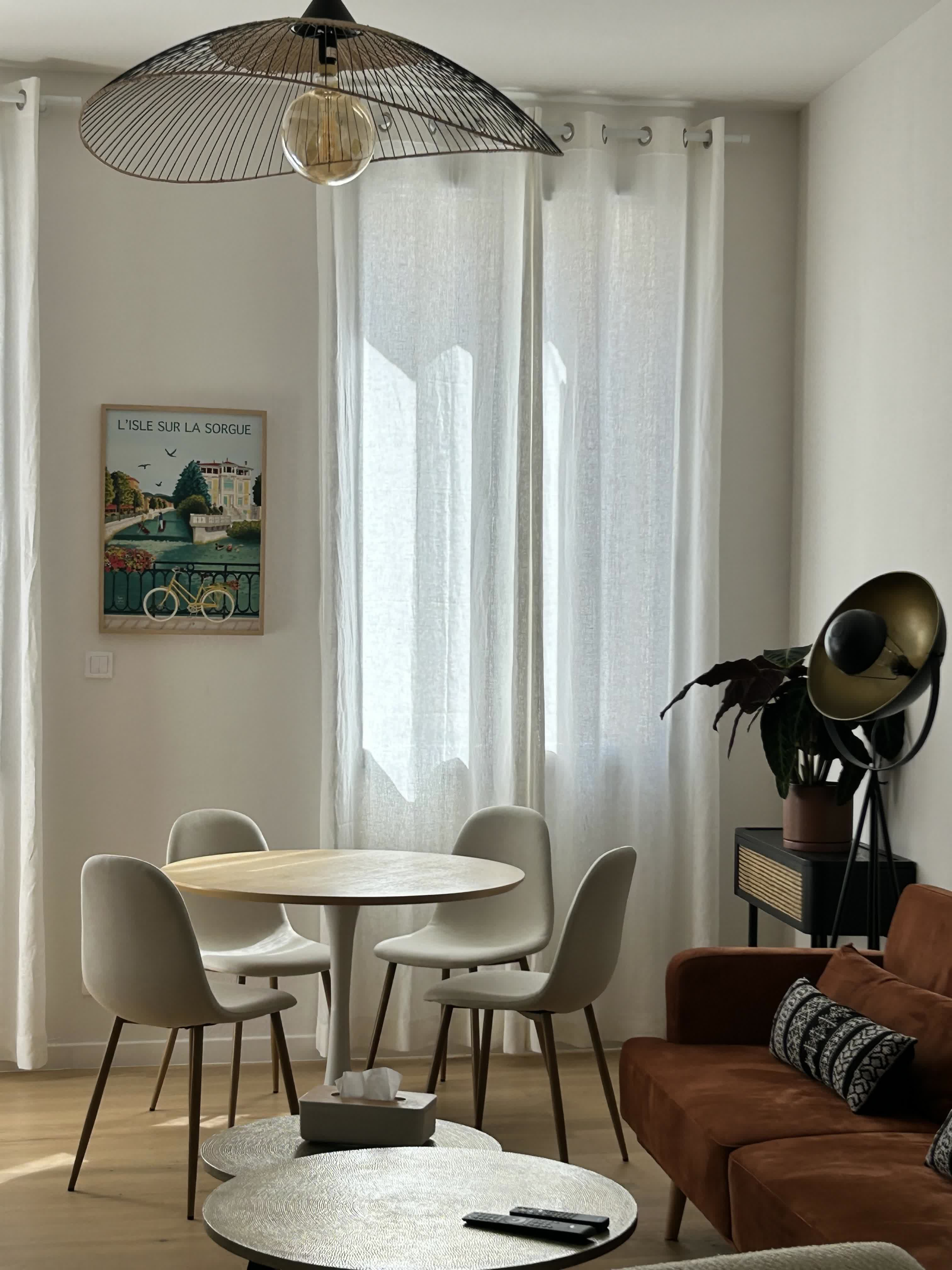 Pièce de vie lumineuse avec un coin repas composé d’une table ronde et de chaises claires, un canapé brun, une grande fenêtre voilée et une décoration moderne aux tons naturels.