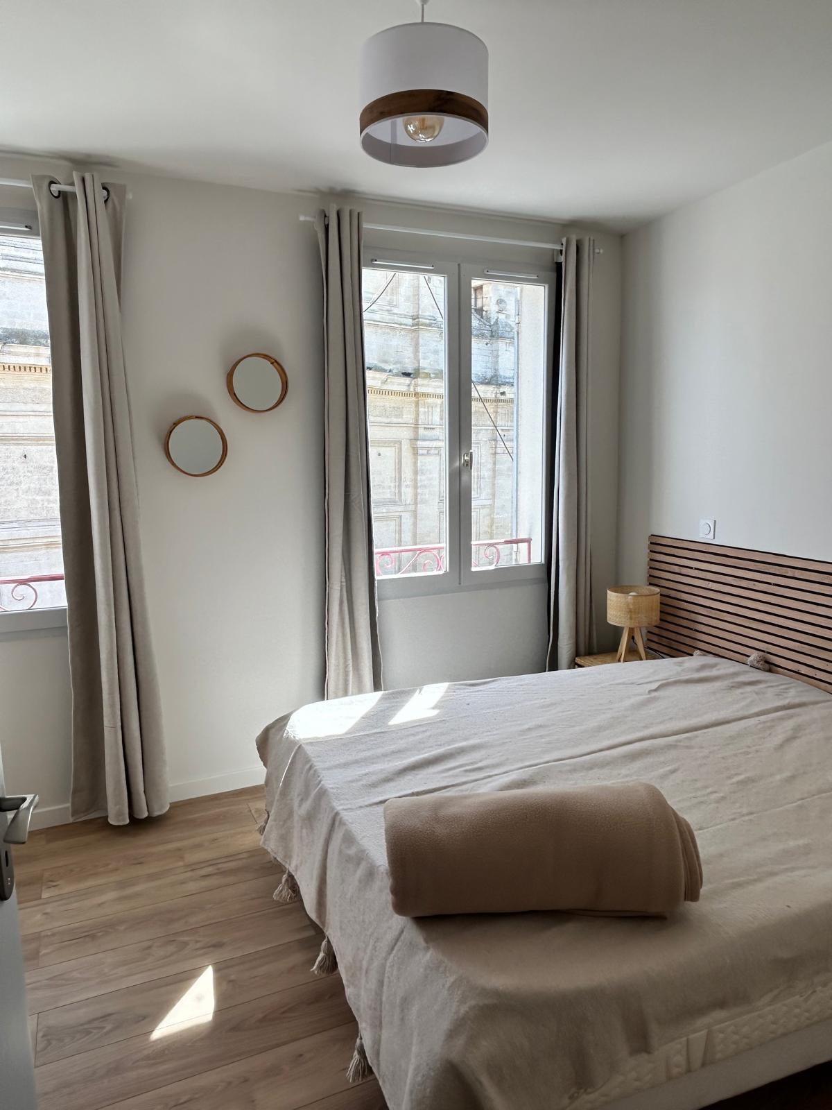 Chambre épurée et lumineuse avec un lit double aux textiles beiges, une tête de lit en bois, des rideaux clairs et une grande fenêtre laissant entrer la lumière du jour.