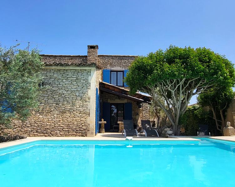 Une maison en pierre de style provençal avec une piscine à débordement et des chaises longues, sous un ciel bleu clair.