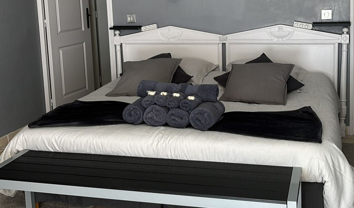 Chambre élégante avec lit double habillé de linge blanc et gris, coussins assortis et serviettes soigneusement disposées.