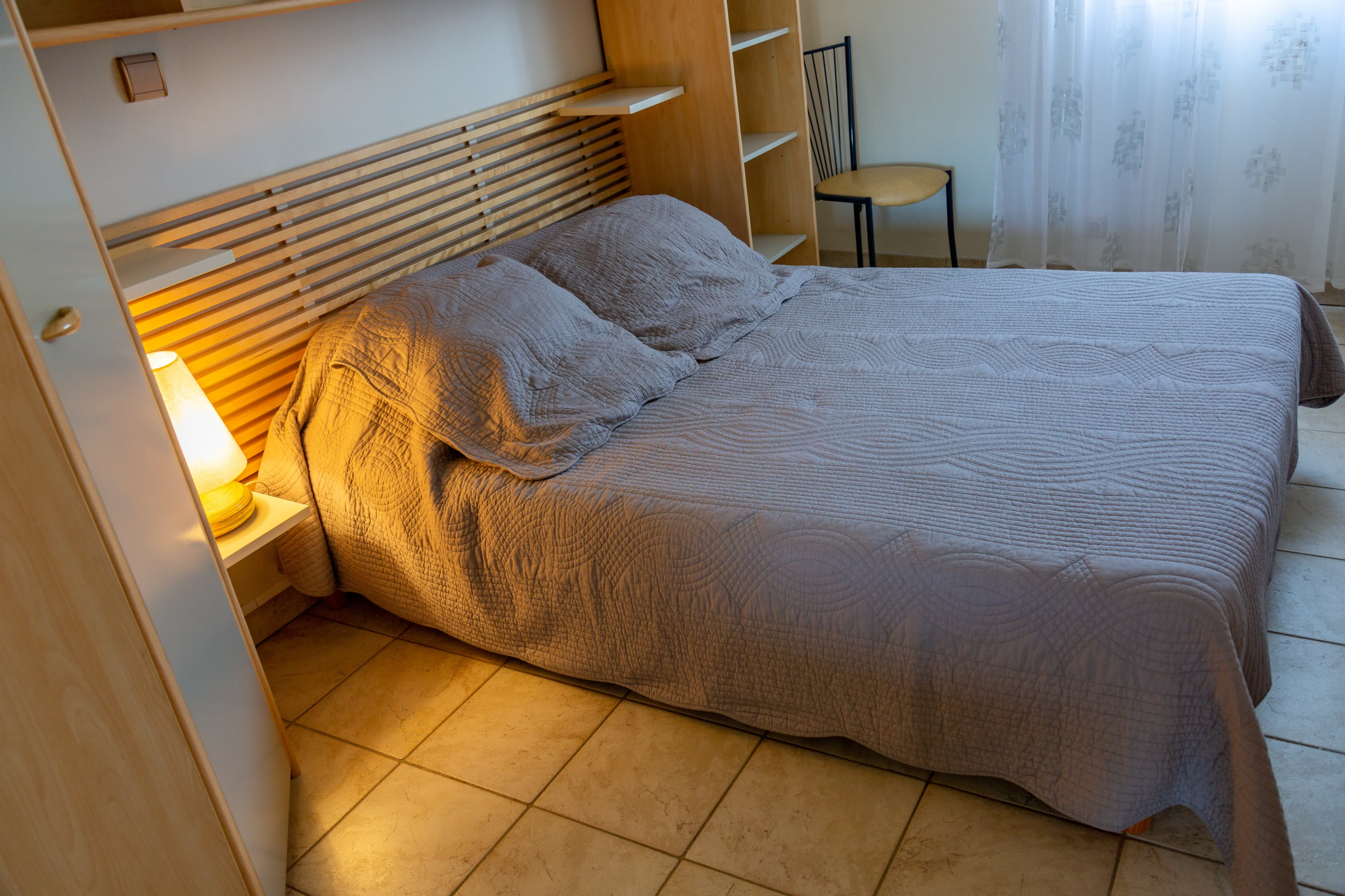Chambre calme avec un lit double aux tons neutres, une tête de lit en bois clair, des rangements intégrés et une lumière douce venant d’une lampe et d’une fenêtre voilée.