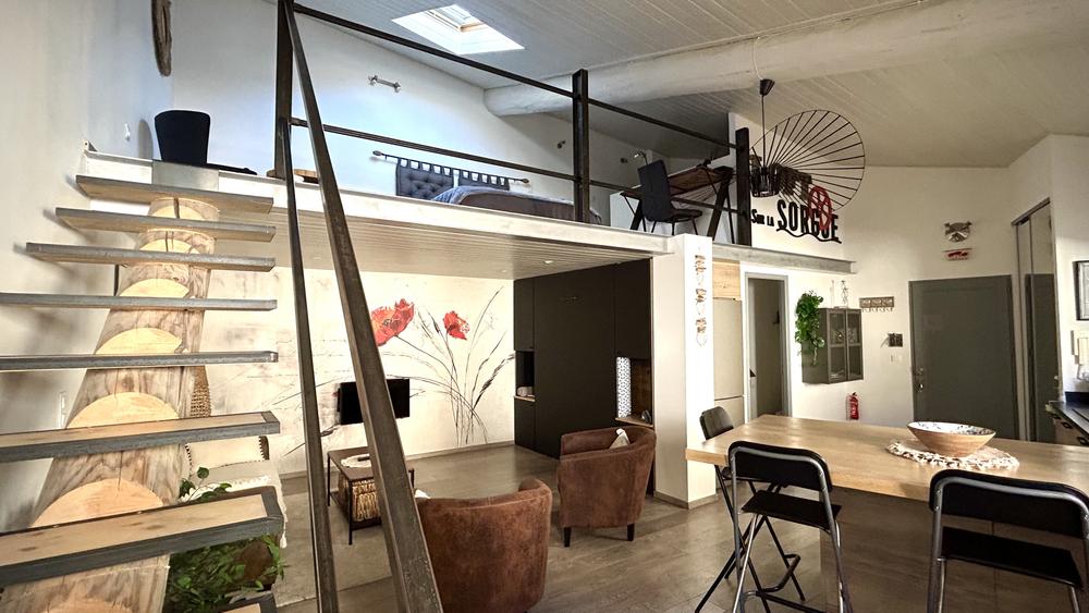 Salon lumineux au style loft avec escalier en bois et métal menant à la mezzanine. Espace convivial composé d’un canapé, de fauteuils et d’une table basse, dans une ambiance moderne et chaleureuse.