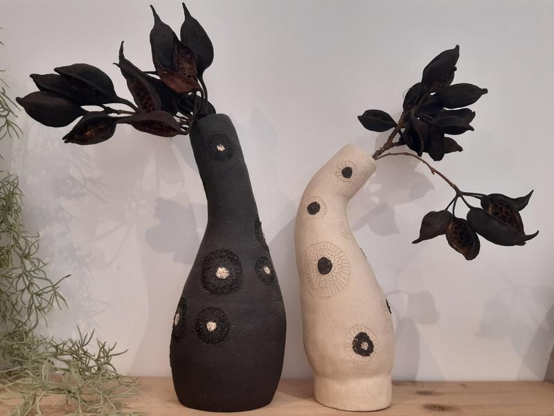 Deux vases sculpturaux en céramique, l’un noir et l’autre beige, aux formes courbes et décorés de motifs circulaires, chacun orné de branches aux feuilles sombres.