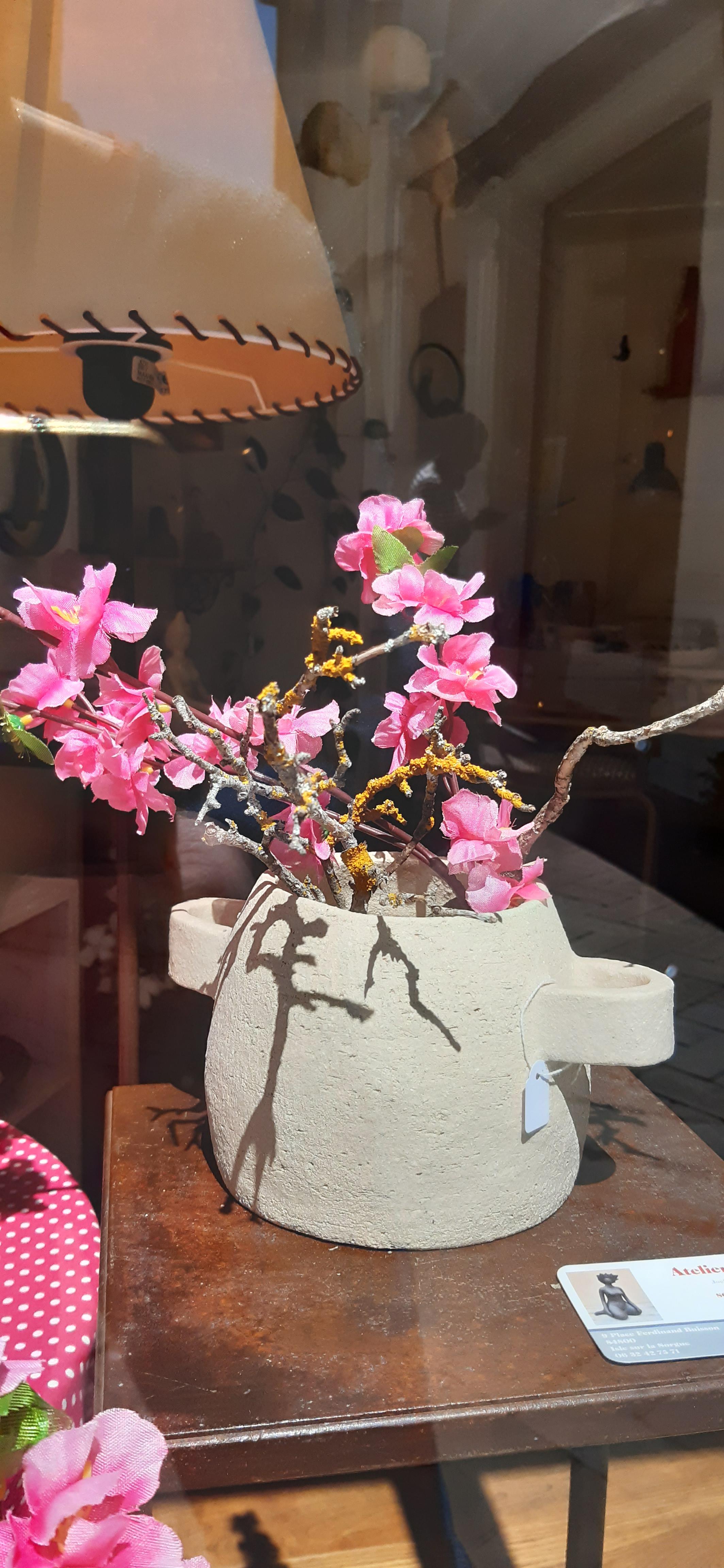 Pot en céramique beige, posé en vitrine, contenant des branches décoratives ornées de fleurs roses vives.
