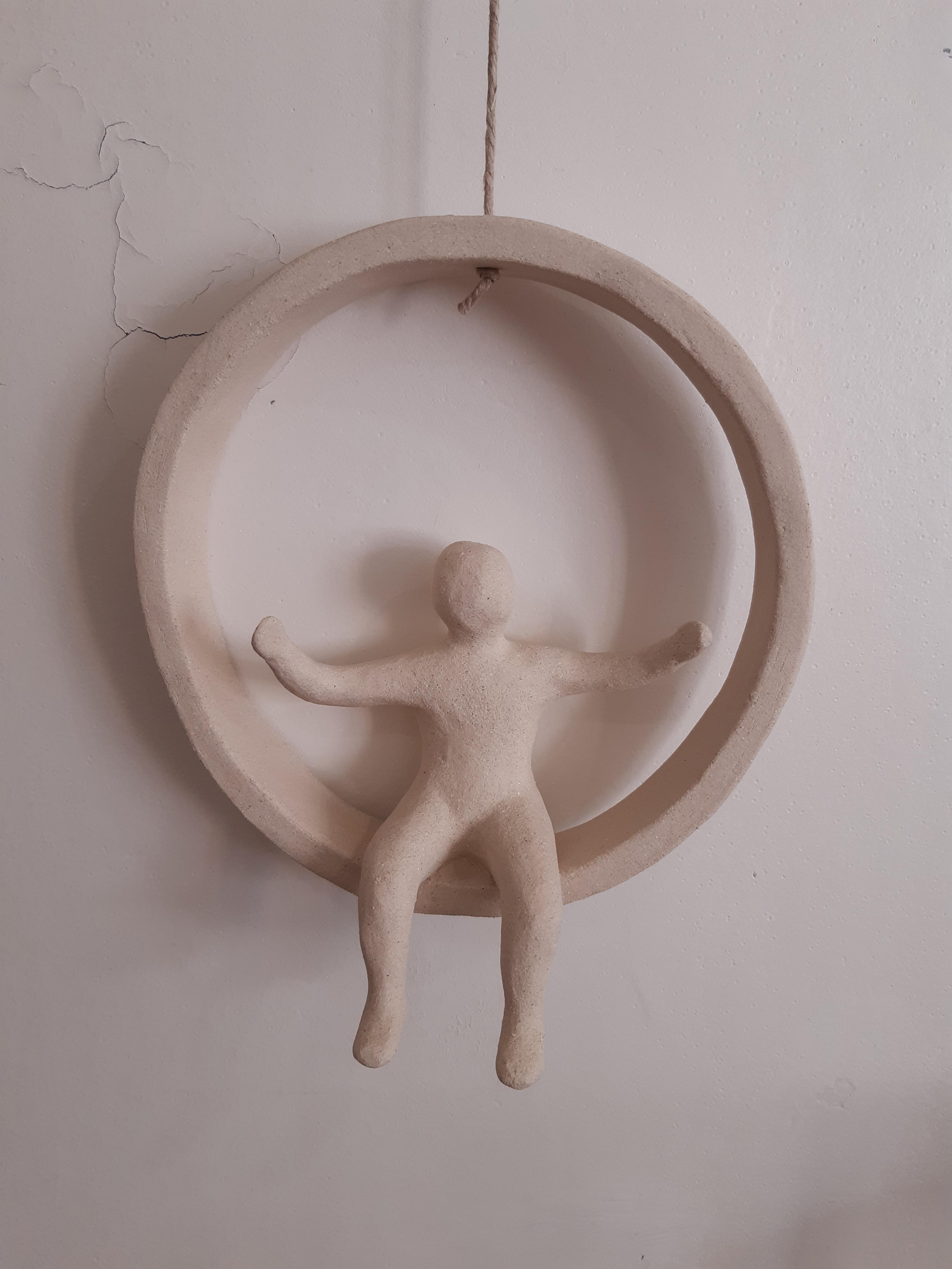 Sculpture murale en céramique beige représentant une silhouette humaine aux bras ouverts, suspendue dans un cercle.