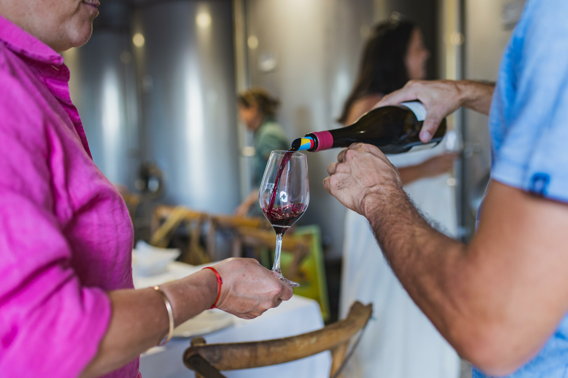 Un homme sert du vin rouge dans un verre tenu par une femme en chemisier rose. La scène, capturée en intérieur avec un arrière-plan flou, évoque une dégustation conviviale dans un domaine viticole.