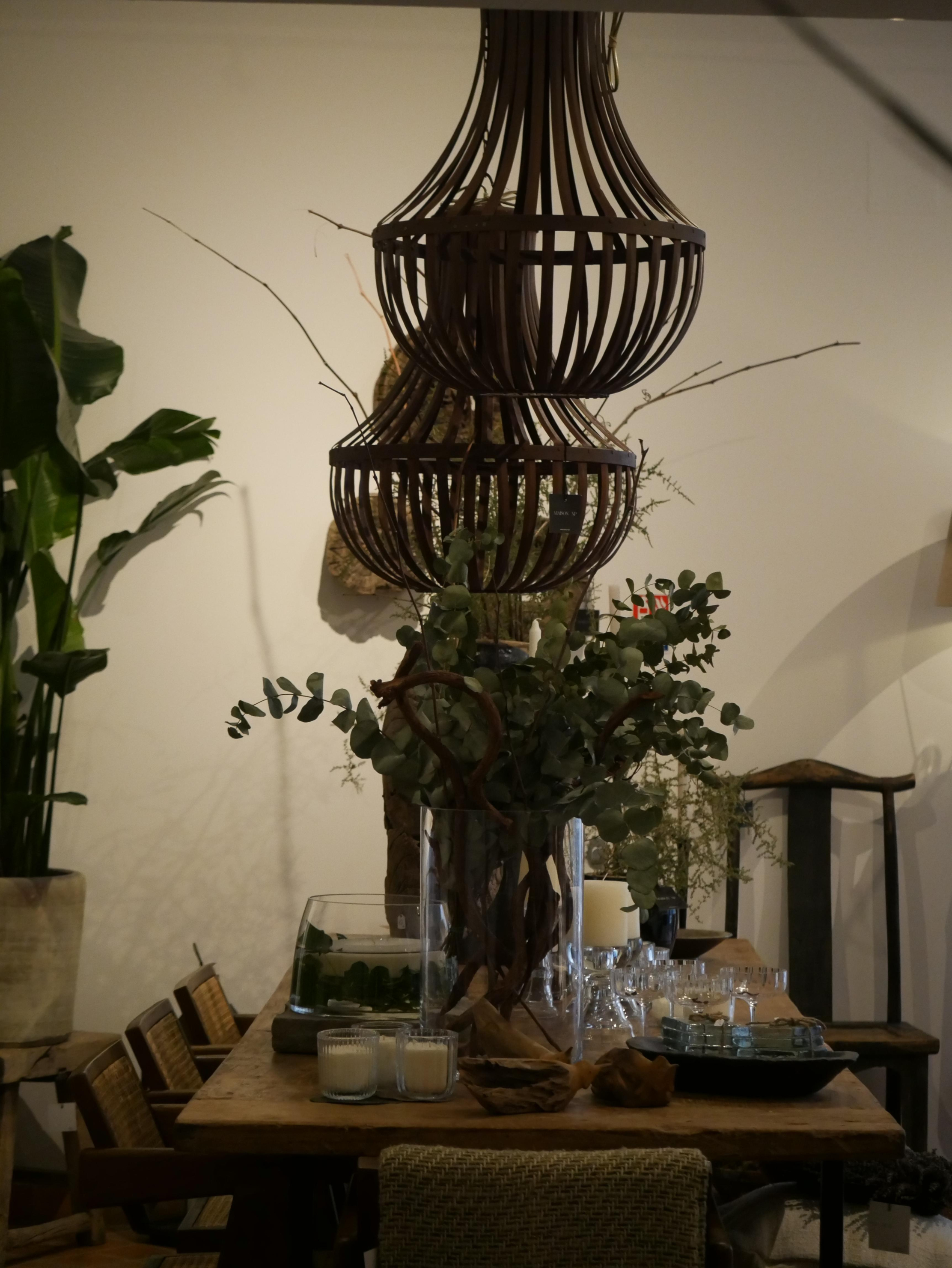 Une grande table de salle à manger en bois est décorée de bougies, de verrerie et d’une imposante composition végétale à base de feuillages et de branches torsadées. Deux grandes suspensions en bois ajouré dominent la scène. L’ensemble crée une atmosphère élégante, organique et intimiste, avec une lumière douce et tamisée.