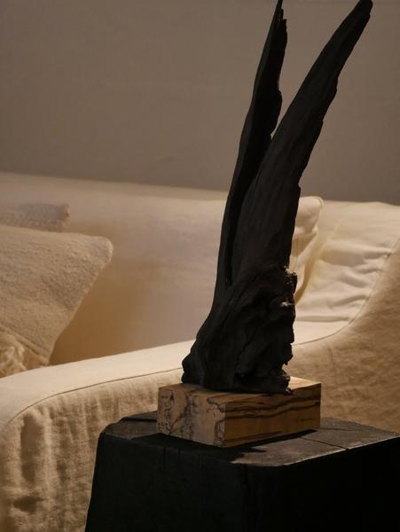 Une sculpture abstraite en bois noirci, élancée et irrégulière, est posée sur un socle en bois clair. Elle se détache devant un canapé blanc aux textiles doux. Le contraste entre le noir de la sculpture et les teintes crème du décor lui donne une présence forte, presque primitive.