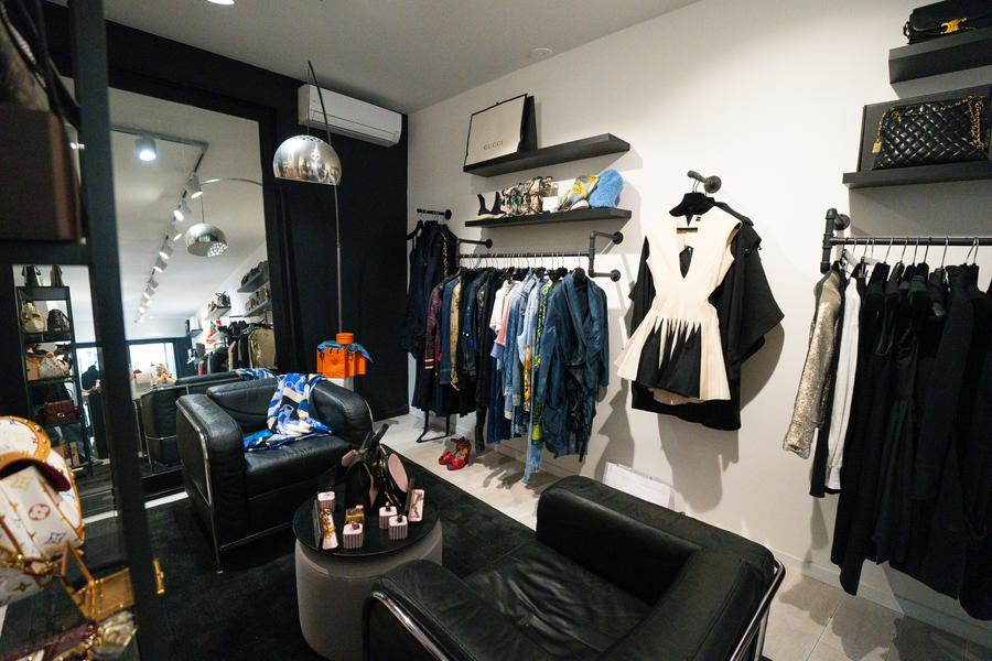 La photo montre l’intérieur d’une boutique de vêtements haut de gamme au style contemporain et soigné.  Au premier plan, on distingue un espace cosy composé de fauteuils en cuir noir disposés autour d’une petite table basse, créant une ambiance presque privée, comme un salon. Sur la gauche, des sacs de grandes marques sont exposés, suggérant une offre orientée luxe ou seconde main premium.  Au fond et sur les murs, plusieurs portants présentent des vêtements soigneusement rangés, principalement dans des tons sobres (noir, blanc, denim). Une robe structurée bicolore attire particulièrement l’attention, mise en avant comme une pièce phare. Des étagères murales accueillent également des accessoires et sacs de créateurs.