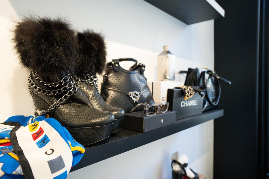 La photo présente une étagère d’exposition mettant en valeur une sélection d’accessoires de mode haut de gamme dans une ambiance élégante et épurée.  Au premier plan, une paire de bottines noires attire immédiatement le regard. Elles se distinguent par leur texture travaillée, leurs chaînes métalliques décoratives et leur finition avec de la fourrure, apportant une touche à la fois audacieuse et sophistiquée. À proximité, on aperçoit un foulard coloré qui contraste avec les tons sombres dominants.  Sur l’étagère, plusieurs sacs à main en cuir noir sont disposés avec soin, accompagnés d’accessoires tels que des lunettes et des objets de petite maroquinerie. Une boîte siglée Chanel est également visible, renforçant l’image d’une sélection issue de grandes maisons de luxe.  L’ensemble est organisé de manière minimaliste, avec une mise en scène soignée qui met en valeur chaque pièce. L’ambiance générale évoque un univers chic et contemporain, typique d’une boutique spécialisée dans les accessoires de créateurs ou le dépôt-vente haut de gamme.