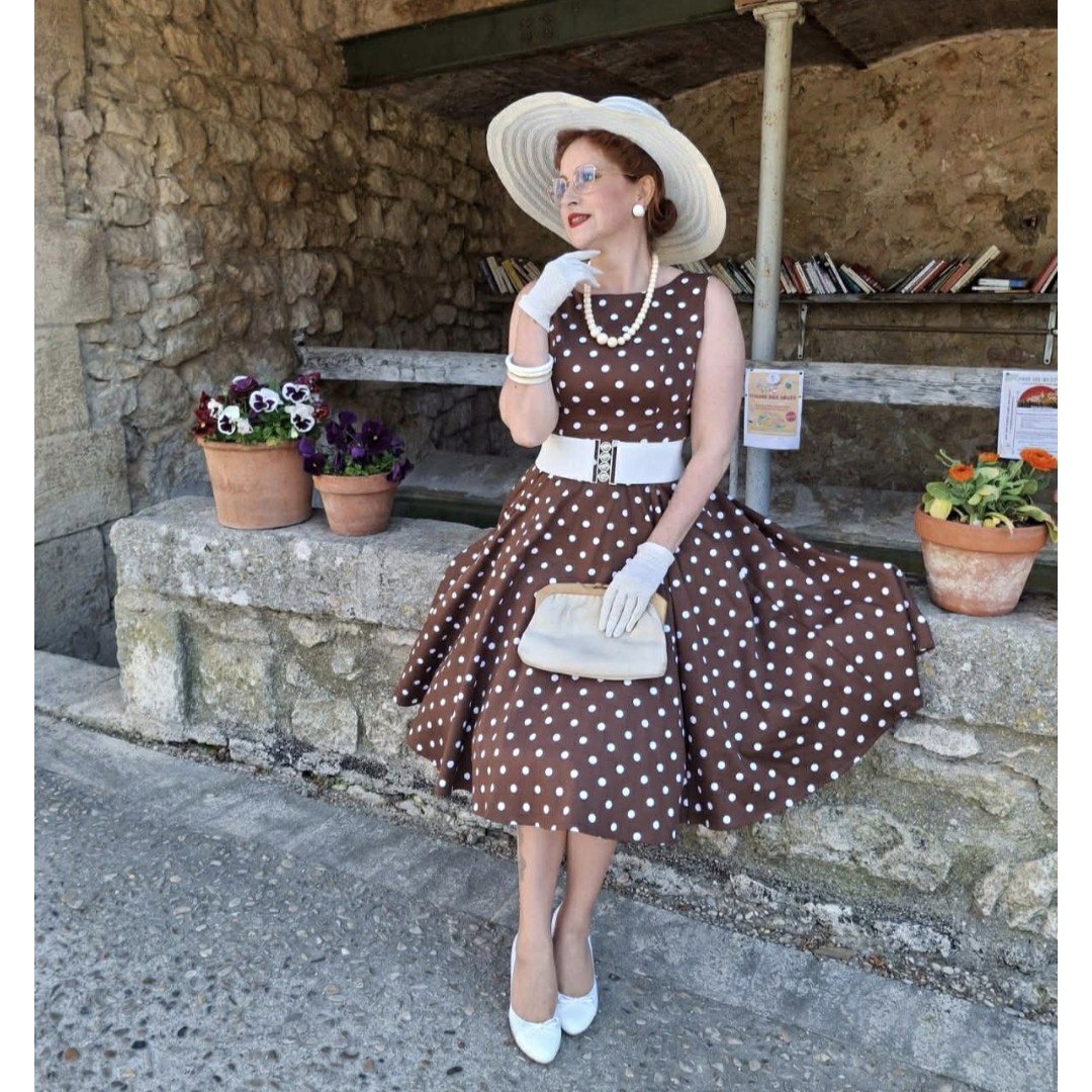 femme assis sur un mur en pierre vêtue d'une robe marron à pois blancs avec chapeau, ceinture large, sac, gants et escarpins blanc