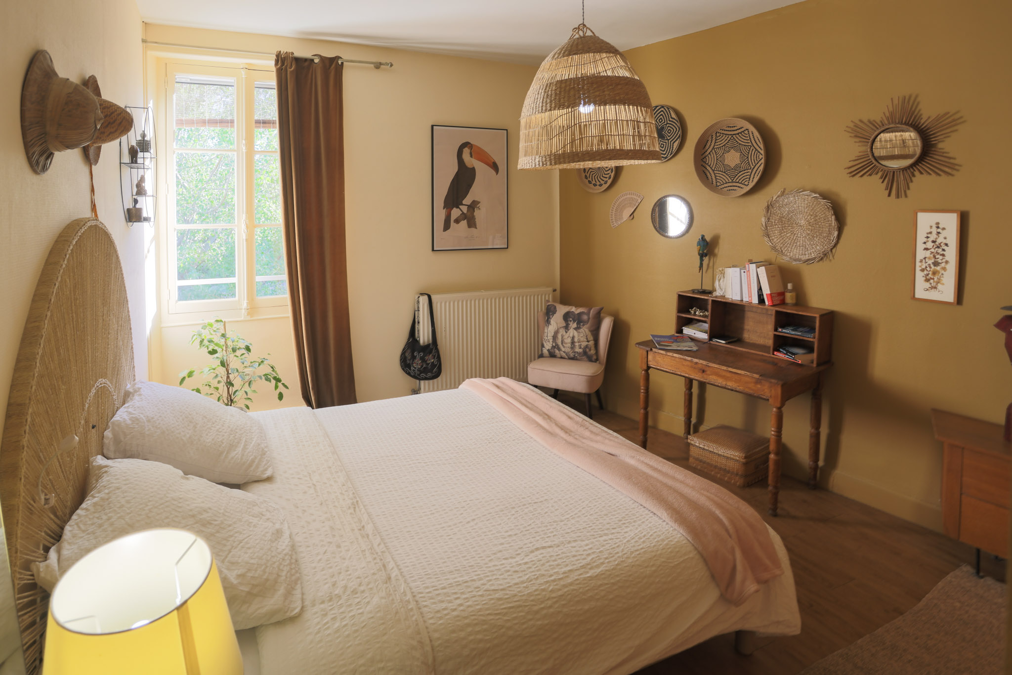 chambre beige avec tete de lit en rotin en demi cercle, au mur miroirs et paniers accrochés. petit bureau ancien en bois
