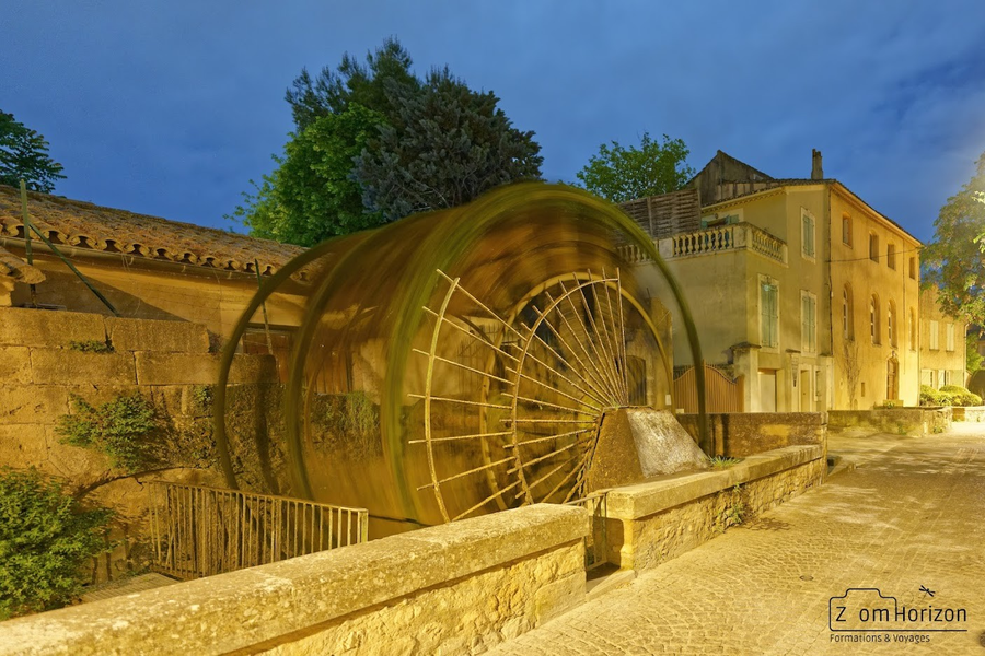 Roue à aubes de L’Isle-sur-la-Sorgue en mouvement, capturée de nuit en pose longue, dans une ambiance chaleureuse entre pierre et lumière.