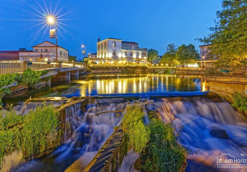 Vue nocturne de L’Isle-sur-la-Sorgue avec une cascade en premier plan, capturée en pose longue. Les lumières des bâtiments se reflètent dans l’eau, créant une ambiance douce et chaleureuse.