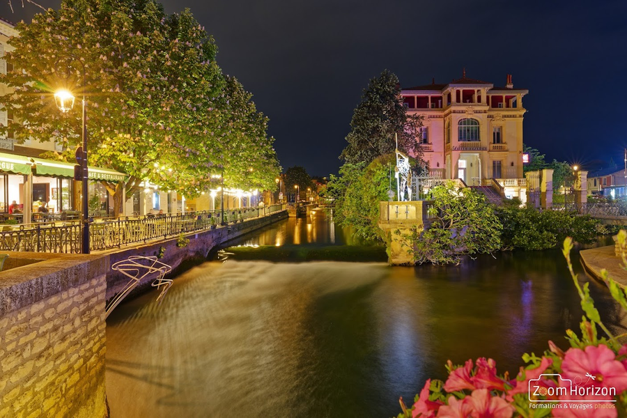 Vue nocturne d’un canal à L’Isle-sur-la-Sorgue, avec une cascade en pose longue et des reflets lumineux. Les façades éclairées et la végétation créent une ambiance chaleureuse et élégante.