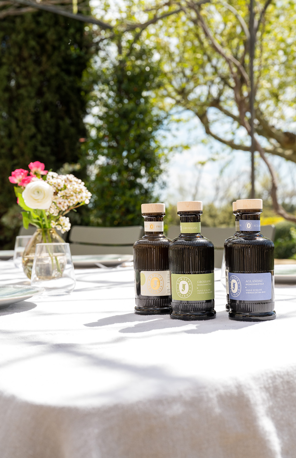 Trois bouteilles d’huile d’olive sont posées sur une grande table recouverte d’une nappe blanche, dans un jardin lumineux. À côté, un petit vase contient quelques fleurs fraîches. En arrière-plan, on devine des arbres et une terrasse ensoleillée. Les bouteilles sont élégantes, sombres, avec des étiquettes colorées et raffinées. L’ensemble donne une impression de repas en plein air, de dégustation soignée et d’art de vivre méditerranéen.
