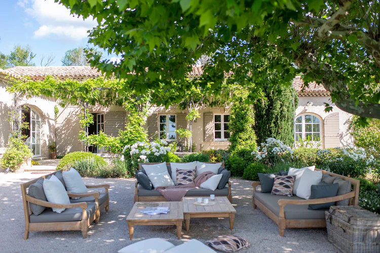 La première image montre une grande terrasse ombragée, aménagée devant une maison de style provençal. Des canapés en bois, garnis de coussins gris et beige, entourent plusieurs tables basses. L’ensemble est encadré par une végétation abondante, des fleurs blanches et de grands arbres, créant une atmosphère paisible et conviviale.