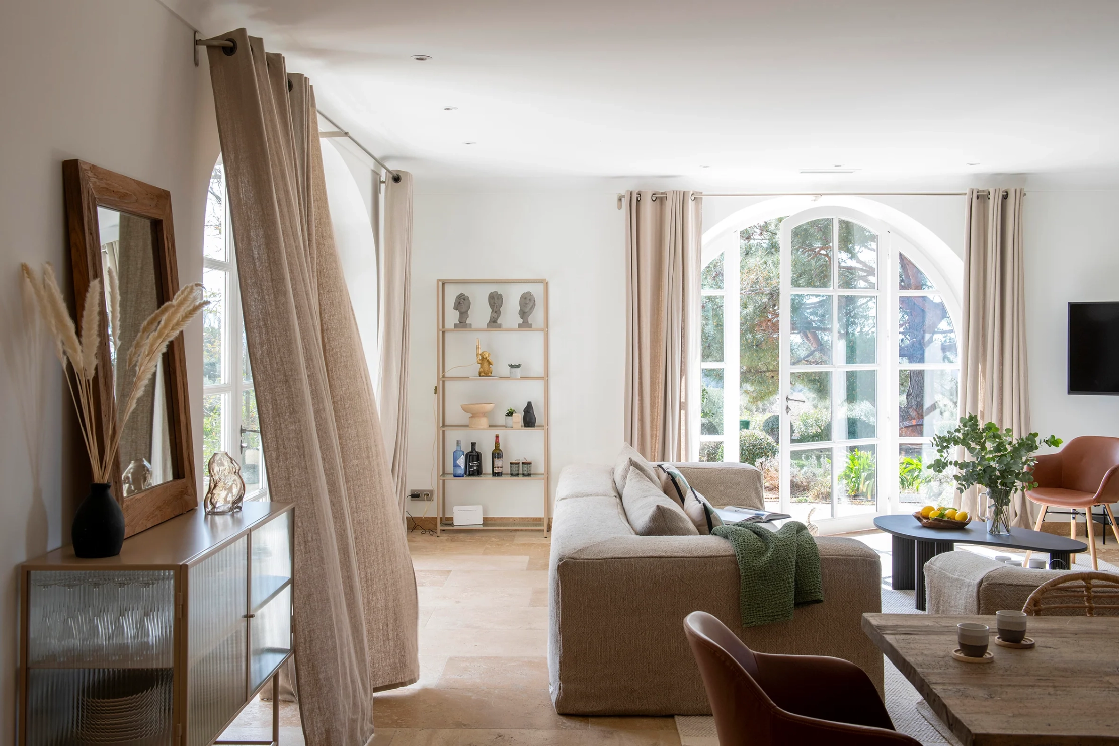 La seconde image présente un vaste séjour lumineux aux tons naturels. De grandes fenêtres cintrées ouvrent sur le jardin. Le mobilier associe un grand canapé beige, des fauteuils en cuir brun, du bois clair et quelques éléments décoratifs minimalistes. Les rideaux en lin, les murs blancs et la pierre au sol renforcent l’ambiance élégante, douce et chaleureuse.