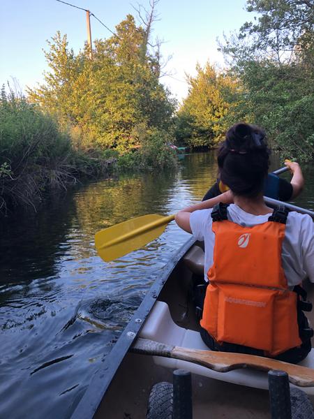 Une personne assise à l’arrière d’un canoë avance doucement sur une rivière calme entourée d’arbres et de hautes herbes. La lumière dorée de la fin de journée se reflète sur l’eau, créant une ambiance paisible et immersive au cœur de la nature.
