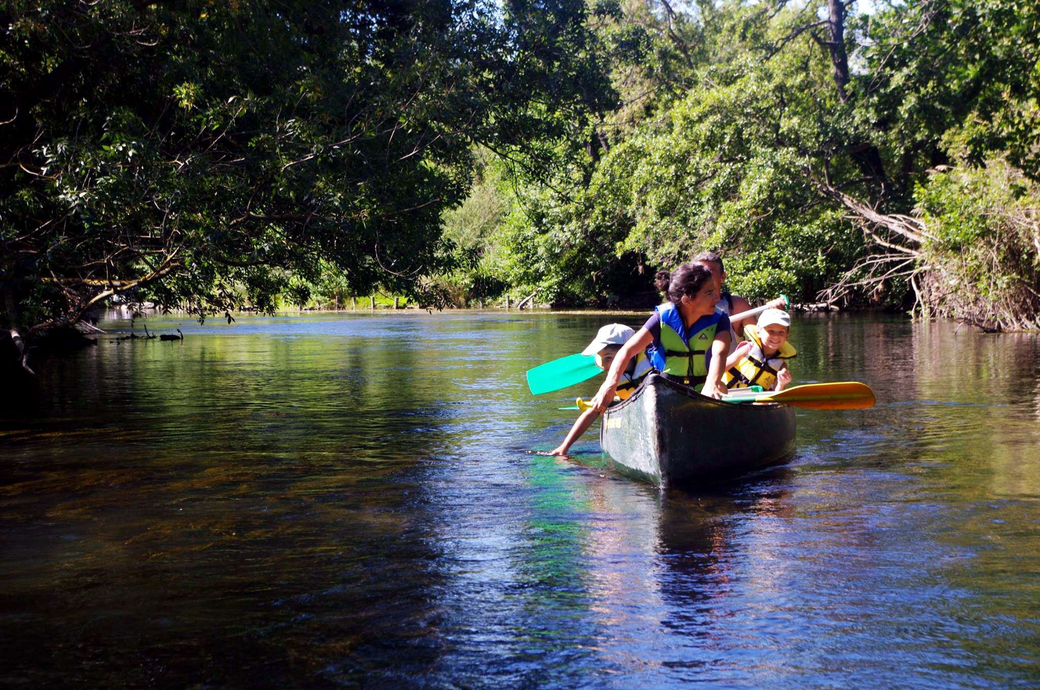 Une famille navigue en canoë sur une rivière aux eaux transparentes bordée d’une végétation dense et verdoyante. Les adultes pagaient tandis que les enfants profitent de la balade dans une atmosphère joyeuse et rafraîchissante.