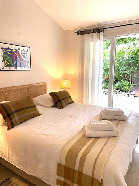 Cette photo présente une chambre élégante et apaisante, baignée de lumière naturelle.  Un grand lit double occupe le centre de la pièce, avec une tête de lit en rotin. Le lit est soigneusement préparé avec une literie blanche et un plaid beige aux tons naturels. Deux coussins décoratifs assortis ajoutent une touche chaleureuse. Des serviettes pliées sont disposées sur le lit, évoquant un hébergement prêt à accueillir des visiteurs. Une lampe de chevet diffuse une lumière douce et chaleureuse. Une grande baie vitrée donne directement sur un petit jardin verdoyant, créant une agréable continuité entre l’intérieur et l’extérieur. Des rideaux légers blancs filtrent la lumière et renforcent l’ambiance douce de la pièce. Un tableau coloré au mur apporte une note artistique et contemporaine.  Cette chambre inspire le repos, la sérénité et le confort.