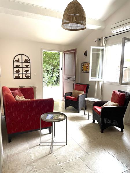 Cette photo montre un salon lumineux et accueillant, aménagé dans un style simple et chaleureux. On y voit :  Un canapé deux places rouge placé à gauche. Deux fauteuils assortis en rotin noir avec coussins rouges, disposés autour de petites tables basses rondes. Un sol carrelé clair qui renforce la luminosité de la pièce. Une porte d’entrée ouverte ainsi qu’une fenêtre ouverte laissant entrer la lumière naturelle et offrant une vue sur la végétation extérieure. Un luminaire suspendu en fibres naturelles au plafond, apportant une touche décorative et chaleureuse. Un cadre mural représentant des silhouettes de chats et une petite œuvre colorée près de la porte. Un climatiseur mural installé au-dessus de la fenêtre.  L’ensemble donne une impression de confort, de calme et de convivialité.