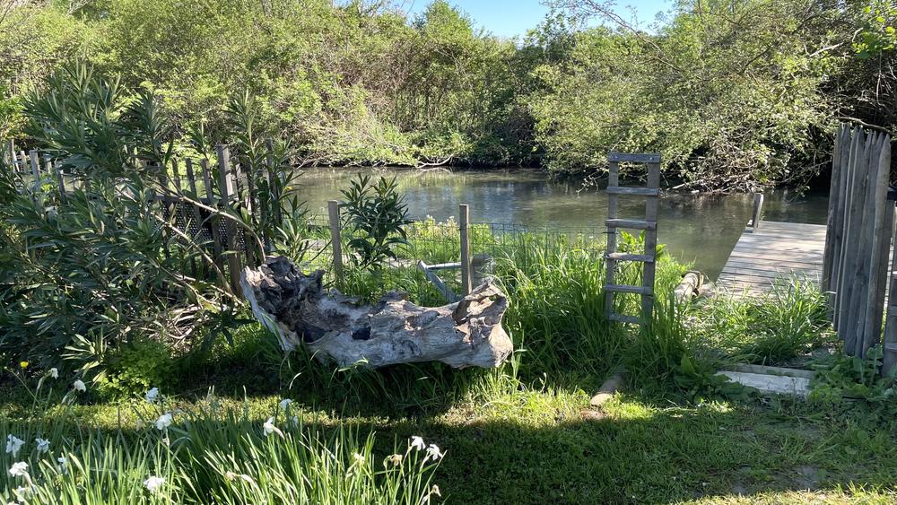 **Photo 4 – Le bord de rivière** Cette photo dévoile un espace naturel particulièrement paisible situé au bord d’un cours d’eau. * Une petite rivière aux eaux calmes serpente au milieu d’une végétation abondante et verdoyante. * Un ponton en bois permet un accès direct à l’eau, offrant un lieu idéal pour contempler le paysage ou profiter d’un moment de détente. * Au premier plan, un imposant tronc d’arbre sculpté par le temps sert d’élément décoratif naturel. * Une échelle en bois rustique et des clôtures discrètes s’intègrent harmonieusement dans l’environnement. * Les berges sont bordées d’herbes, de fleurs et d’arbustes qui renforcent le caractère sauvage et authentique du lieu. * Les arbres environnants se reflètent dans l’eau et créent une agréable sensation de fraîcheur et d’intimité. * La lumière du soleil met en valeur les différentes nuances de vert et souligne la beauté naturelle du site. Cet espace offre un cadre bucolique et ressourçant, propice à la détente, à la contemplation de la nature et à l’évasion au fil de l’eau.