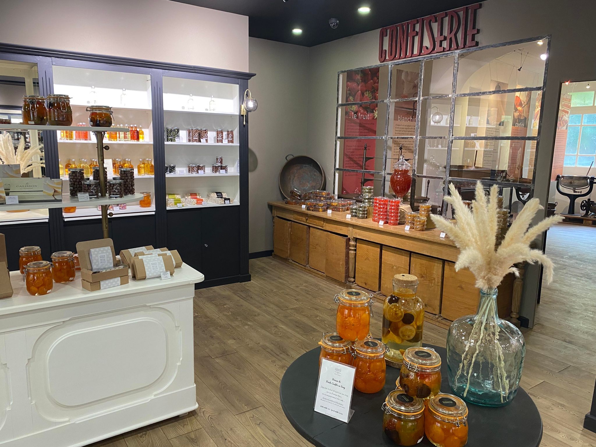 Boutique élégante et chaleureuse de confiserie artisanale. La pièce est lumineuse, avec un parquet clair et des meubles mêlant bois ancien et vitrines modernes noires et blanches. Des bocaux de fruits confits et de sirops colorés sont exposés sur plusieurs tables et étagères. Au fond, une grande inscription « Confiserie » domine un espace rappelant un ancien atelier. L’ambiance évoque à la fois la tradition provençale et une présentation contemporaine haut de gamme.