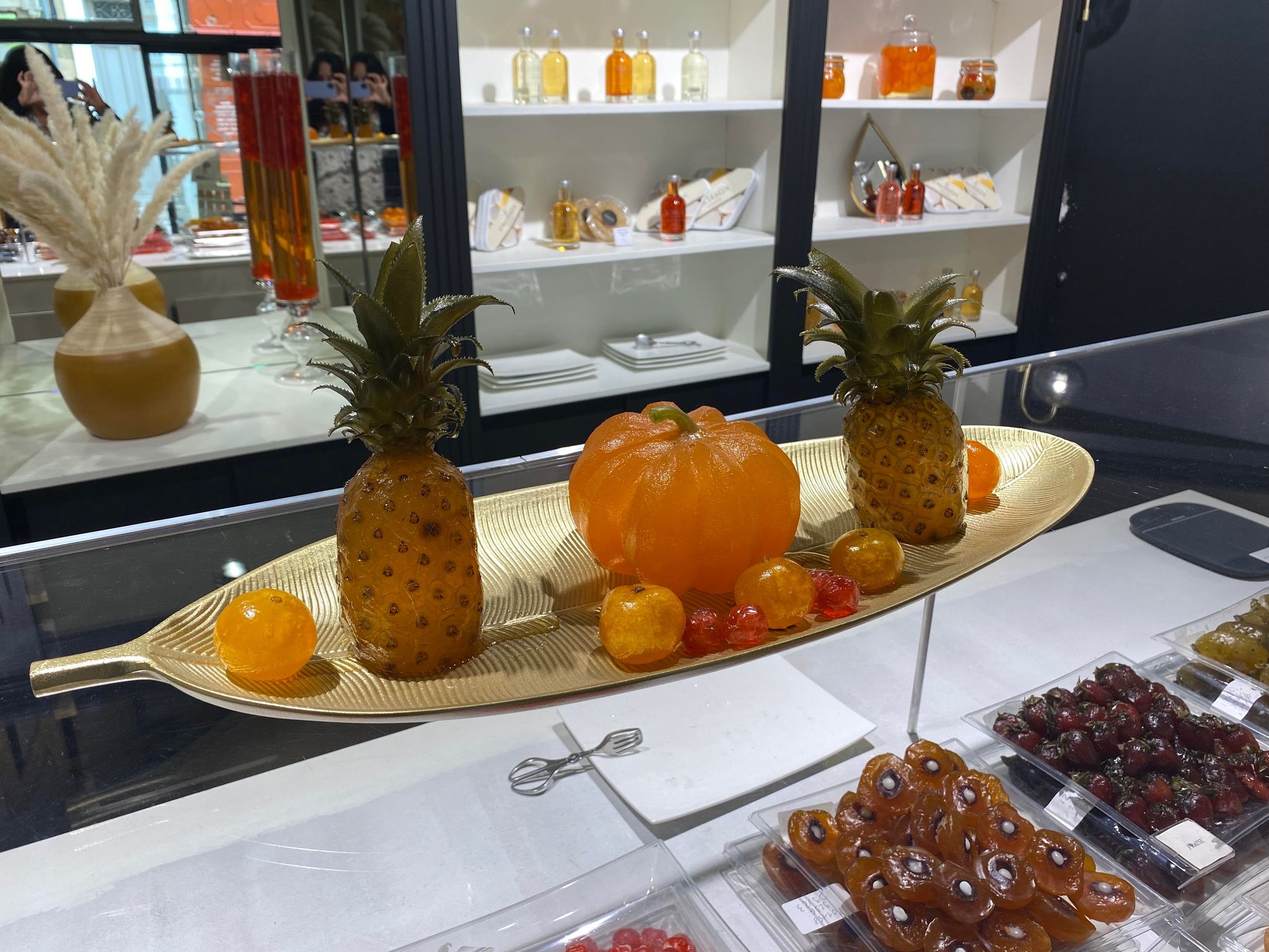 Gros plan sur une présentation raffinée de fruits confits. Deux ananas confits et une petite courge orange translucide sont disposés sur un plateau doré en forme de feuille. Autour, plusieurs fruits confits colorés brillent sous la lumière. En arrière-plan, des étagères blanches exposent des coffrets et bouteilles de sirops dans une atmosphère chic et gourmande.