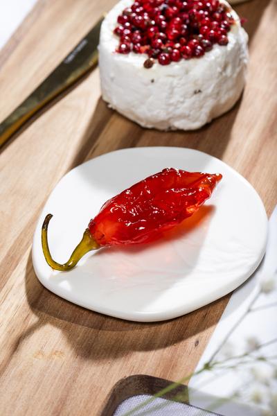 Un piment rouge confit, brillant et translucide, est présenté sur une petite assiette blanche. À l’arrière, un fromage de chèvre frais couvert de baies roses repose sur une planche en bois. La scène met en valeur une association sucrée-salée originale et gastronomique, dans une ambiance culinaire raffinée.