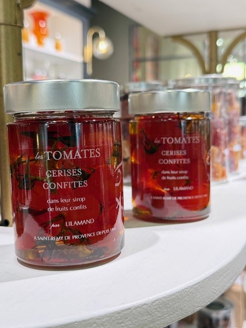 Deux pots en verre de tomates cerises confites sont posés sur une étagère blanche. Les tomates rouges brillent dans leur sirop transparent. Les étiquettes rouges et noires mettent en avant le savoir-faire artisanal de la maison Lilamand. L’arrière-plan est flou, laissant apparaître une boutique élégante et lumineuse.