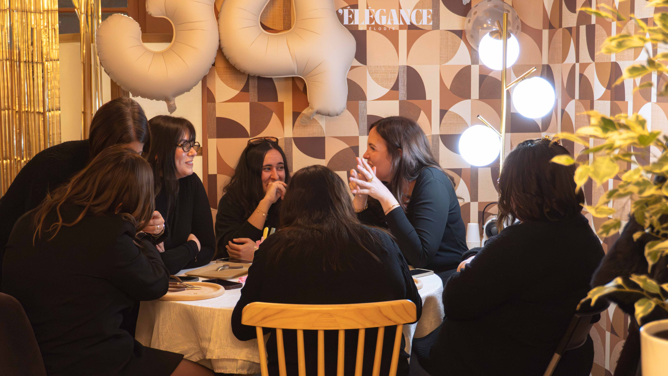 Un groupe de femmes est réuni autour d’une table dans une ambiance chaleureuse et intimiste. Elles discutent et participent à un atelier créatif de bijoux sous une lumière douce, avec une atmosphère conviviale et détendue.