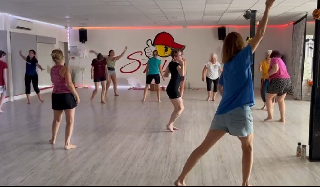 Cette seconde photo met en scène un moment d’exploration corporelle collective au sein d’une séance de danse libre intuitive. Les participants occupent l’ensemble de la salle et adoptent des postures variées, témoignant de la liberté de mouvement offerte par la pratique. Au premier plan, une participante lève les bras dans un geste ample et spontané, tandis que les autres explorent différentes dynamiques corporelles. L’image traduit une atmosphère bienveillante et énergisante, propice à l’expression de soi, à la créativité et au lâcher-prise.